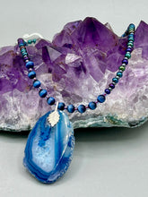 Load image into Gallery viewer, This Vibrant Royal Blue Agate Geode Sliced Stone Chip Necklace is a bold and beautiful statement piece. It features a natural gemstone pendant in a blend of light and dark blue tones, surrounded by glass beads in rich hues of blue, purple, gold, and green. This chic, boho-inspired necklace is completed by 10 blue cats eye beads and a silver leaf bail.  Measurements:   Necklace is 18” in length - pendant is 2 3/4” Height
