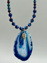 Load image into Gallery viewer, This Vibrant Royal Blue Agate Geode Sliced Stone Chip Necklace is a bold and beautiful statement piece. It features a natural gemstone pendant in a blend of light and dark blue tones, surrounded by glass beads in rich hues of blue, purple, gold, and green. This chic, boho-inspired necklace is completed by 10 blue cats eye beads and a silver leaf bail.  Measurements:   Necklace is 18” in length - pendant is 2 3/4” Height
