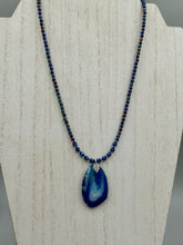 Load image into Gallery viewer, This Vibrant Royal Blue Agate Geode Sliced Stone Chip Necklace is a bold and beautiful statement piece. It features a natural gemstone pendant in a blend of light and dark blue tones, surrounded by glass beads in rich hues of blue, purple, gold, and green. This chic, boho-inspired necklace is completed by 10 blue cats eye beads and a silver leaf bail.  Measurements:   Necklace is 18” in length - pendant is 2 3/4” Height
