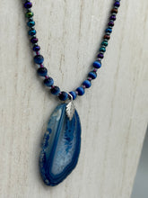 Load image into Gallery viewer, This Vibrant Royal Blue Agate Geode Sliced Stone Chip Necklace is a bold and beautiful statement piece. It features a natural gemstone pendant in a blend of light and dark blue tones, surrounded by glass beads in rich hues of blue, purple, gold, and green. This chic, boho-inspired necklace is completed by 10 blue cats eye beads and a silver leaf bail.  Measurements:   Necklace is 18” in length - pendant is 2 3/4” Height
