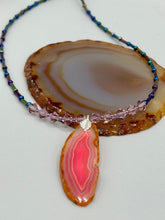 Load image into Gallery viewer, Make a bold style statement with this eye-catching Soft Pink and Amber Agate Geode Sliced Stone Chip Necklace. Featuring a vibrant soft pink and amber tone agate geode pendant surrounded by dark blue glass beads with iridescent tones of purple, blue, green, pink and gold. This bohemian-style gemstone necklace is the perfect way to add a unique touch of shabby boho chic to any look.   Measurements:    Necklace is 18” in length - pendant is 2 1/4” Height
