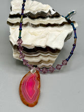Load image into Gallery viewer, Make a bold style statement with this eye-catching Soft Pink and Amber Agate Geode Sliced Stone Chip Necklace. Featuring a vibrant soft pink and amber tone agate geode pendant surrounded by dark blue glass beads with iridescent tones of purple, blue, green, pink and gold. This bohemian-style gemstone necklace is the perfect way to add a unique touch of shabby boho chic to any look.   Measurements:    Necklace is 18” in length - pendant is 2 1/4” Height
