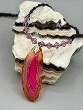 Load image into Gallery viewer, Make a bold style statement with this eye-catching Soft Pink and Amber Agate Geode Sliced Stone Chip Necklace. Featuring a vibrant soft pink and amber tone agate geode pendant surrounded by dark blue glass beads with iridescent tones of purple, blue, green, pink and gold. This bohemian-style gemstone necklace is the perfect way to add a unique touch of shabby boho chic to any look.   Measurements:    Necklace is 18” in length - pendant is 2 1/4” Height
