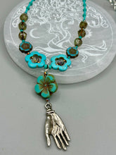 Load image into Gallery viewer, Adorn your look and show your affinity for nature with this exquisite handcrafted necklace, composed of a large intricate sea-green hibiscus pressed glass bead, a lucky hand charm, multiple turquoise, and dark avocado green faceted pressed glass beads, and a hook-eye clasp for secure wear. This nature-inspired statement piece brings warmth to any outfit for both day and night. Measurements:  Necklace: 19 1/2&quot;, Flower and lucky hand pendant 3&quot;
