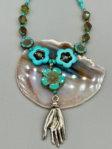 Adorn your look and show your affinity for nature with this exquisite handcrafted necklace, composed of a large intricate sea-green hibiscus pressed glass bead, a lucky hand charm, multiple turquoise, and dark avocado green faceted pressed glass beads, and a hook-eye clasp for secure wear. This nature-inspired statement piece brings warmth to any outfit for both day and night. Measurements:  Necklace: 19 1/2