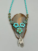 Load image into Gallery viewer, Adorn your look and show your affinity for nature with this exquisite handcrafted necklace, composed of a large intricate sea-green hibiscus pressed glass bead, a lucky hand charm, multiple turquoise, and dark avocado green faceted pressed glass beads, and a hook-eye clasp for secure wear. This nature-inspired statement piece brings warmth to any outfit for both day and night. Measurements:  Necklace: 19 1/2&quot;, Flower and lucky hand pendant 3&quot;
