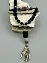 Load image into Gallery viewer, This elegant black obsidian square-shaped beaded necklace is the perfect accessory for your unique look. With a large black bead adorned with multi-color cloisonné bead and a lucky hand charm, it exudes a dark, alluring vibe. Complemented with black glass and hematite stone beads, it will make a bold statement.  Measurements:  Necklace: 18” in length - lucky hand charm is 3 1 /2” Height
