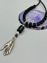 Load image into Gallery viewer, This elegant black obsidian square-shaped beaded necklace is the perfect accessory for your unique look. With a large black bead adorned with multi-color cloisonné bead and a lucky hand charm, it exudes a dark, alluring vibe. Complemented with black glass and hematite stone beads, it will make a bold statement.  Measurements:  Necklace: 18” in length - lucky hand charm is 3 1 /2” Height
