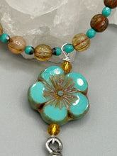 Load image into Gallery viewer, Complete your look with this carefully crafted necklace, featuring a large sea-green hibiscus pressed glass bead, a lucky hand charm, assorted champagne melon beads, and turquoise glass beads. All these elements are combined with a hook-eye clasp, ensuring lasting wear. This nature-inspired accessory adds a touch of charm to all kinds of outfits, making it ideal for both day and night. Measurements:   Necklace: 18&quot;
