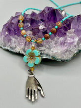 Load image into Gallery viewer, Complete your look with this carefully crafted necklace, featuring a large sea-green hibiscus pressed glass bead, a lucky hand charm, assorted champagne melon beads, and turquoise glass beads. All these elements are combined with a hook-eye clasp, ensuring lasting wear. This nature-inspired accessory adds a touch of charm to all kinds of outfits, making it ideal for both day and night. Measurements:   Necklace: 18&quot;
