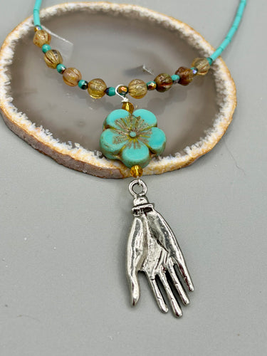 Complete your look with this carefully crafted necklace, featuring a large sea-green hibiscus pressed glass bead, a lucky hand charm, assorted champagne melon beads, and turquoise glass beads. All these elements are combined with a hook-eye clasp, ensuring lasting wear. This nature-inspired accessory adds a touch of charm to all kinds of outfits, making it ideal for both day and night. Measurements:   Necklace: 18