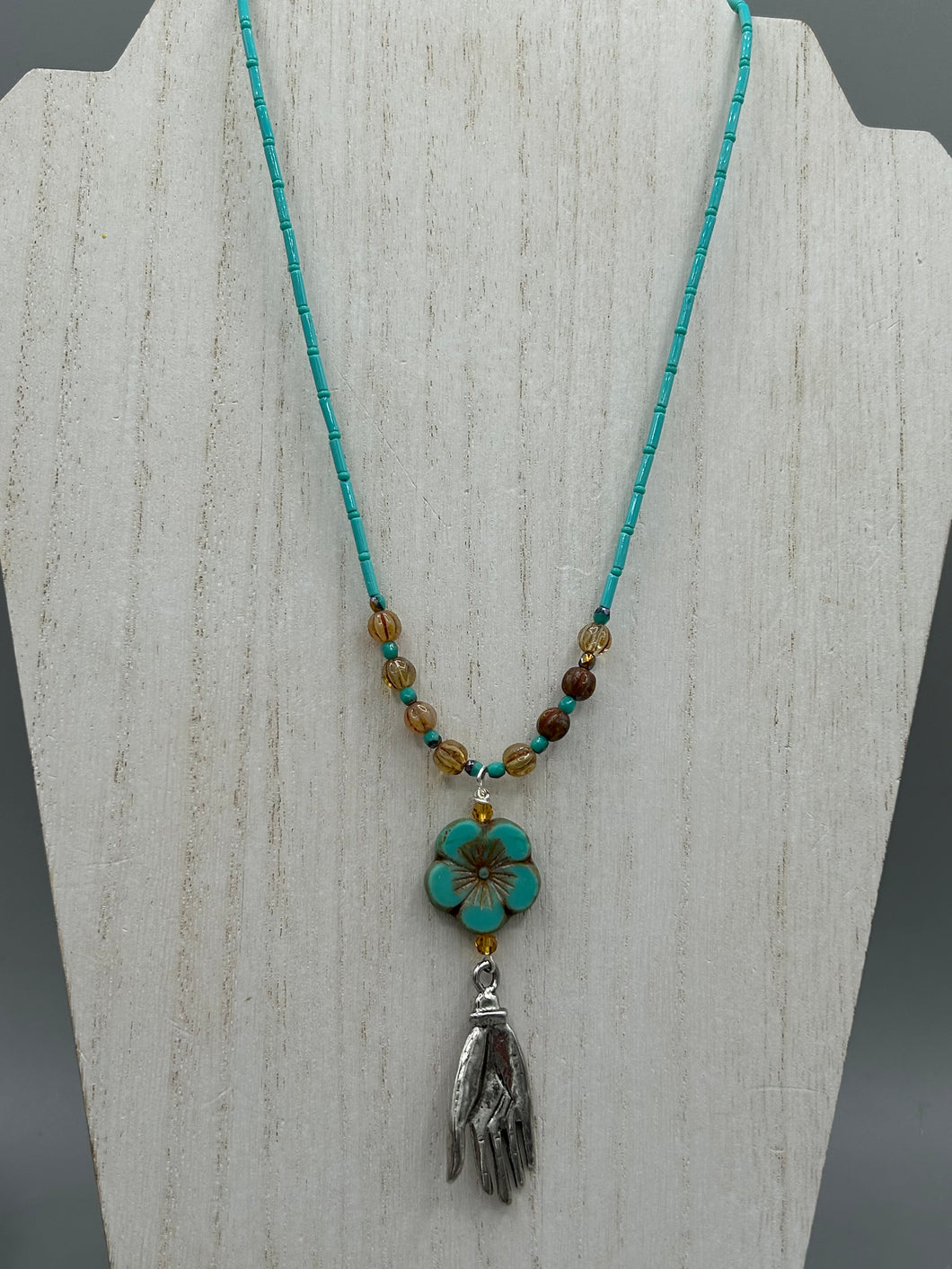 Complete your look with this carefully crafted necklace, featuring a large sea-green hibiscus pressed glass bead, a lucky hand charm, assorted champagne melon beads, and turquoise glass beads. All these elements are combined with a hook-eye clasp, ensuring lasting wear. This nature-inspired accessory adds a touch of charm to all kinds of outfits, making it ideal for both day and night. Measurements:   Necklace: 18