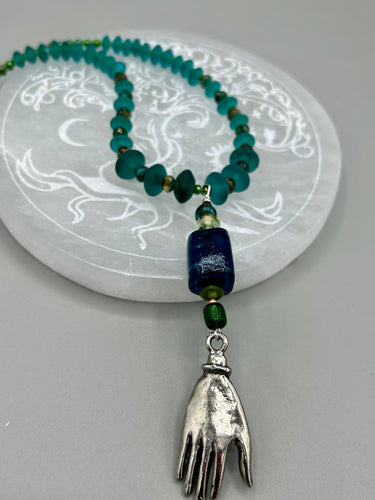 Bring the beauty of nature to your wardrobe with this gorgeous blue-green glass beaded necklace. Featuring a large rectangular blue-green glass bead, a lucky hand charm, mixed flat matte teal glass beads, and a hook-eye clasp for secure wear, this nature-inspired accessory adds a touch of summer to any outfit. Perfect for day-to-night looks.   Measurements:  Necklace: 19