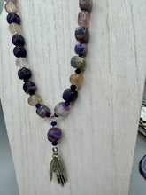 Load image into Gallery viewer,  This nature-inspired accessory features an array of unique hues, including rainbow fluorite stone beads and iridescent dark shimmery purple glass beads with hints of gold, blue, pink, and green. The secure hook-eye clasp makes it perfect for day to night wear, allowing you to add a touch of summer to your wardrobe. The necklace is further complemented by a large square-shaped rainbow fluorite bead and a lucky hand charm. Measurements:  Necklace: 18&quot;, Lucky hand pendant 3&quot;
