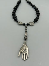 Load image into Gallery viewer, This sophisticated onyx beaded necklace features a large lucky hand charm adorned with floral metal beads, conveying an enchanting aura. The piece is finished with long black glass and floral metal beaded hanging strands, creating a bold and engaging visual statement. Measurements:  Necklace: 19” in length - lucky hand charm is 4” Height
