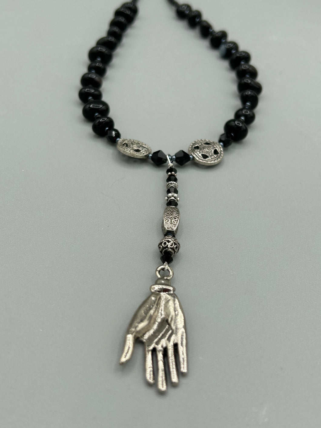 This sophisticated onyx beaded necklace features a large lucky hand charm adorned with floral metal beads, conveying an enchanting aura. The piece is finished with long black glass and floral metal beaded hanging strands, creating a bold and engaging visual statement. Measurements:  Necklace: 19” in length - lucky hand charm is 4” Height