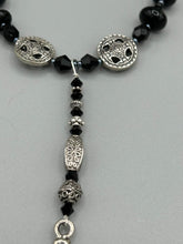 Load image into Gallery viewer, This sophisticated onyx beaded necklace features a large lucky hand charm adorned with floral metal beads, conveying an enchanting aura. The piece is finished with long black glass and floral metal beaded hanging strands, creating a bold and engaging visual statement. Measurements:  Necklace: 19” in length - lucky hand charm is 4” Height
