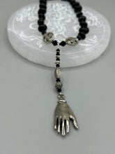 Load image into Gallery viewer, This sophisticated onyx beaded necklace features a large lucky hand charm adorned with floral metal beads, conveying an enchanting aura. The piece is finished with long black glass and floral metal beaded hanging strands, creating a bold and engaging visual statement. Measurements:  Necklace: 19” in length - lucky hand charm is 4” Height
