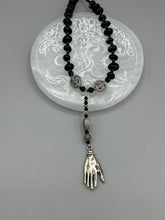 Load image into Gallery viewer, This sophisticated onyx beaded necklace features a large lucky hand charm adorned with floral metal beads, conveying an enchanting aura. The piece is finished with long black glass and floral metal beaded hanging strands, creating a bold and engaging visual statement. Measurements:  Necklace: 19” in length - lucky hand charm is 4” Height

