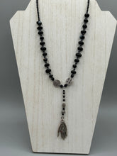 Load image into Gallery viewer, This sophisticated onyx beaded necklace features a large lucky hand charm adorned with floral metal beads, conveying an enchanting aura. The piece is finished with long black glass and floral metal beaded hanging strands, creating a bold and engaging visual statement. Measurements:  Necklace: 19” in length - lucky hand charm is 4” Height
