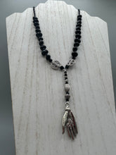 Load image into Gallery viewer, This sophisticated onyx beaded necklace features a large lucky hand charm adorned with floral metal beads, conveying an enchanting aura. The piece is finished with long black glass and floral metal beaded hanging strands, creating a bold and engaging visual statement. Measurements:  Necklace: 19” in length - lucky hand charm is 4” Height
