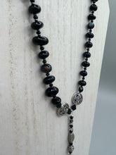 Load image into Gallery viewer, This sophisticated onyx beaded necklace features a large lucky hand charm adorned with floral metal beads, conveying an enchanting aura. The piece is finished with long black glass and floral metal beaded hanging strands, creating a bold and engaging visual statement. Measurements:  Necklace: 19” in length - lucky hand charm is 4” Height

