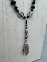 Load image into Gallery viewer, This sophisticated onyx beaded necklace features a large lucky hand charm adorned with floral metal beads, conveying an enchanting aura. The piece is finished with long black glass and floral metal beaded hanging strands, creating a bold and engaging visual statement. Measurements:  Necklace: 19” in length - lucky hand charm is 4” Height
