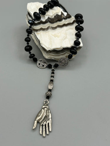 This sophisticated onyx beaded necklace features a large lucky hand charm adorned with floral metal beads, conveying an enchanting aura. The piece is finished with long black glass and floral metal beaded hanging strands, creating a bold and engaging visual statement. Measurements:  Necklace: 19” in length - lucky hand charm is 4” Height