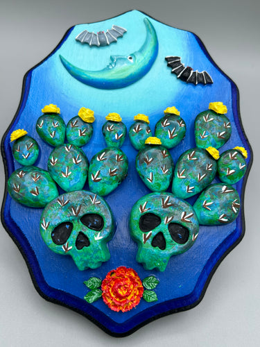 This carefully crafted polymer clay wall art showcases a wood frame adorned with a crescent moon motif. A striking color palette of deep sky tones, including light blue and royal blue to black, further enhances the piece. Delicate cacti plants bloom with light yellow flowers, with spooky skulls are incorporated among the cacti plant and a finishing touch of flying bat motifs.  Measurements: H 9