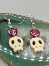Load image into Gallery viewer, These stylish skull earrings are perfect for adding a unique flair to any outfit. Whether you&#39;re looking for a Dia De Los Muertos accessory or simply looking to draw inspiration, these goth pieces perfectly capture the vampire goth, witchy, dark, Chingona Cabrona, Latina Chicana styles. Howlite stone skull beaded earrings come with a ruby red heart-shaped glass beaded that has a very shimmery and vivid iridescent hue.  Measurements: 1 1/4 ” in length
