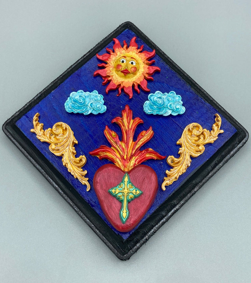 Handcrafted polymer clay wall art is displayed on a square wood frame, featuring sun and light blue cloud motifs on each side and a dark royal blue backdrop with a bright red sacred heart in the center. A turquoise and yellow cross provides contrast to the warm toned heart, which is further embellished with gold accents on each side of the heart.