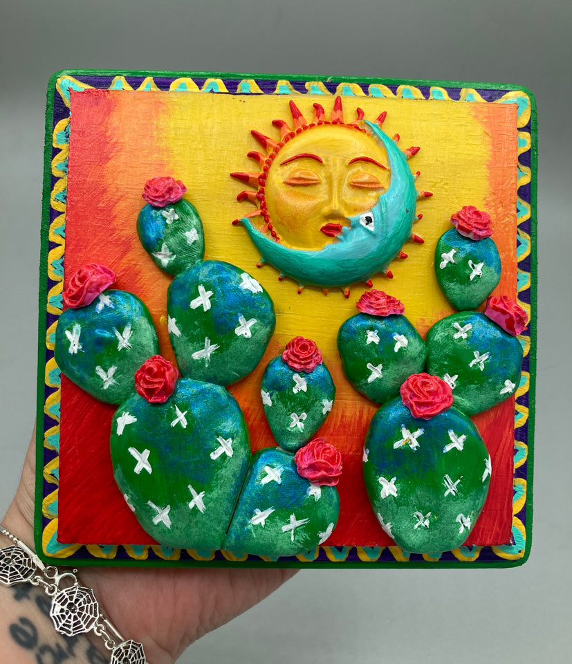 Handcrafted polymer clay wall art on a square-shaped wood frame with a sun and crescent moon motifs. Its warm sky hues, including yellow, orange and dark red, are further complemented by hand sculpted cacti plants with cool green-blue tones and dark pink flowers. The dark green frame adds an extra layer of sophistication to the warm-toned artwork.