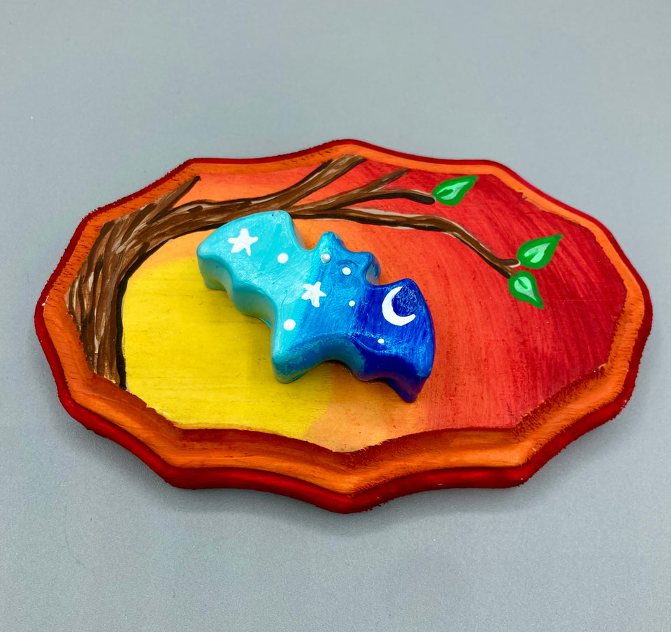 This unique Starry Night Mini Bat Wall Art Decor is hand sculpted and painted in vivid hues to offer an eye-catching decoration for your living space. The bat is painted in a gradient of light to dark blue, and is complemented by white stars and a crescent moon. The wood frame is painted in yellow, orange and red for a