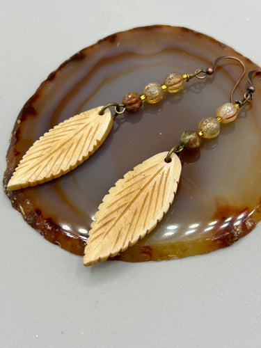 Single-stranded earring features bone carved leaf charm earrings with champagne mix melon Czech faceted round glass beads.  Glass beads have a tint of light champagne, amber, dark green and gold shimmer. Antique Copper Plated Surgical Steel Hammered Long Ear-wire French Hook Earrings.   Measurements: 3” in length  