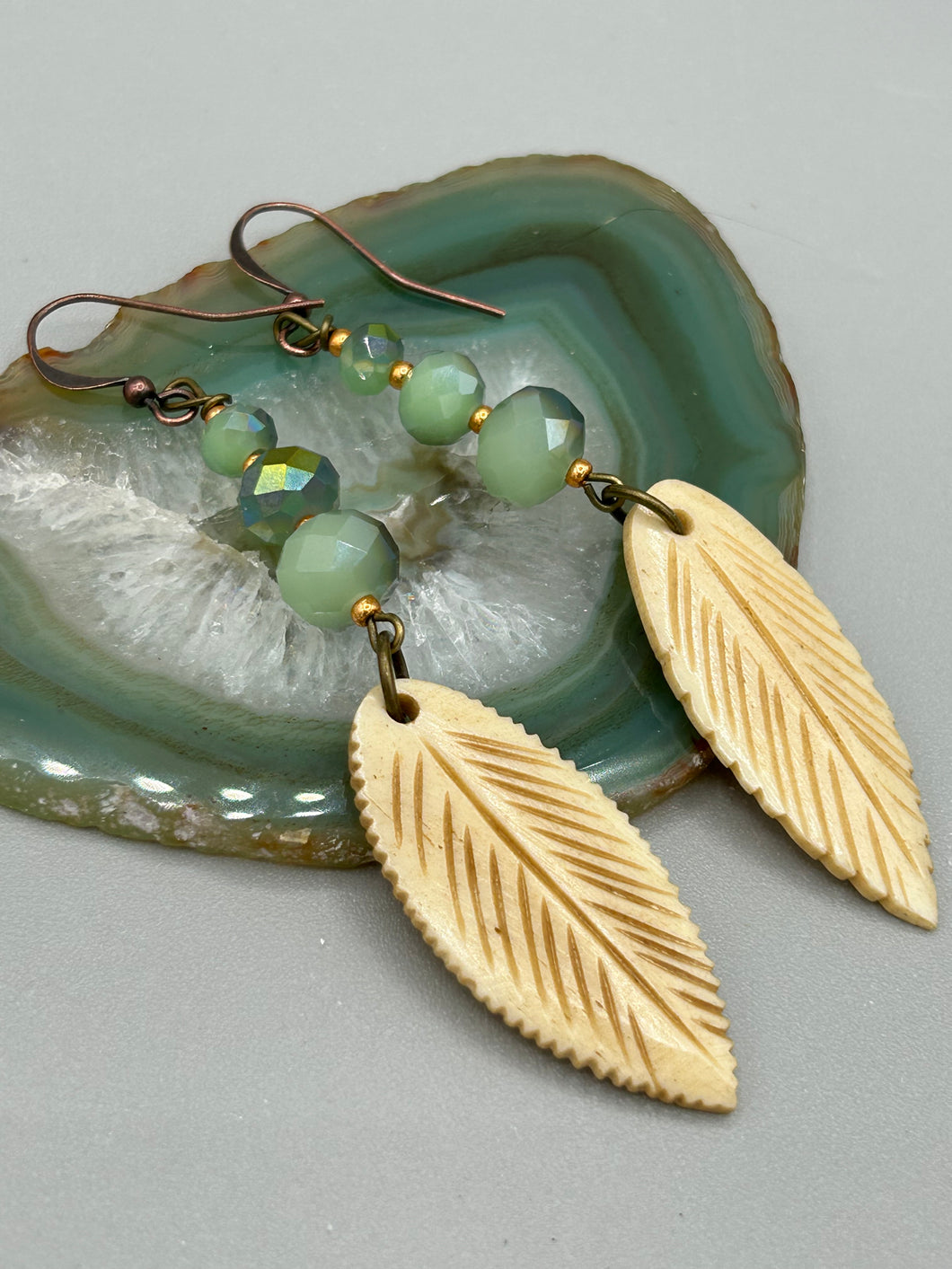 Single-stranded earring features bone carved leaf charm earrings with light mint faceted round glass beads.  Glass beads have a tint of dark green and gold shimmer. Antique Copper Plated Surgical Steel Hammered Long Ear-wire French Hook Earrings.  Measurements: 3” in length  