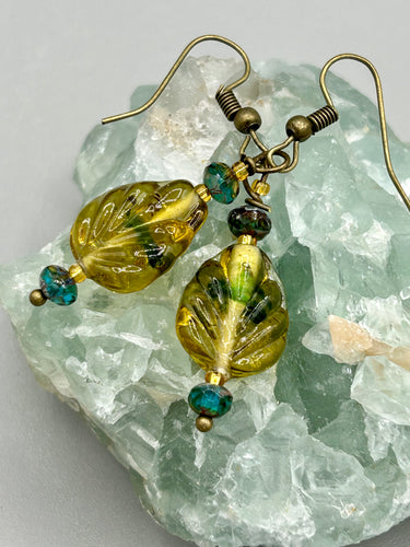 This pair of earrings features a dark amber and green tone, pressed glass leaf charm in an iridescent finish that gives off subtle hues of gold. Complemented with dark teal and gold glass beaded earrings, this set is sure to sparkle.  Measurements: 1 1/4