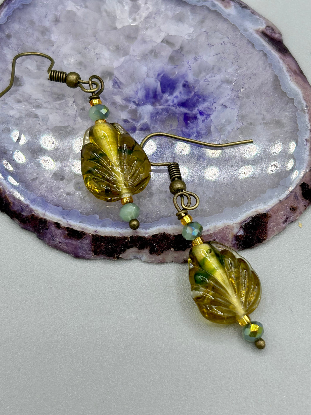 This pair of earrings features a dark amber and green tone, pressed glass leaf charm in an iridescent finish that gives off subtle hues of gold. Complemented with light ming green glass beaded earrings, this set is sure to sparkle.   Measurements: 1 1/4