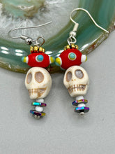 Load image into Gallery viewer, Spooky but festive skull beads with hematite bead spines and come with fiesta shaped crown glass earrings. These stylish skull earrings are perfect for adding a unique flair to any outfit. Whether you&#39;re looking for a Dia De Los Muertos accessory or simply looking to draw inspiration, these goth pieces perfectly capture the vampire goth, witchy, dark, Chingona Cabrona, Latina Chicana styles.   Measurements: 1 1/2” in length
