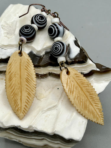 Single-stranded earring features bone carved leaf charm earrings with black and white swirly eye-dot glass beads. Antique Copper Plated Surgical Steel Hammered Long Ear-wire French Hook Earrings. Measurements: 3” in length  