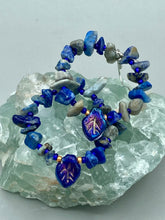 Load image into Gallery viewer, These Lapis Stone Chip Beaded Hoop Earrings feature a dark blue-tone pressed glass leaf charm with an iridescent shimmer finish showcasing hues of gold, purple, blue and pink. The earrings display natural lapis stone chips and royal blue with gold glass beads.  Measurements: 1 1/2&quot; length
