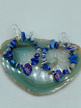 Load image into Gallery viewer, These Lapis Stone Chip Beaded Hoop Earrings feature a dark blue-tone pressed glass leaf charm with an iridescent shimmer finish showcasing hues of gold, purple, blue and pink. The earrings display natural lapis stone chips and royal blue with gold glass beads.  Measurements: 1 1/2&quot; length
