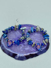 Load image into Gallery viewer, These Lapis Stone Chip Beaded Hoop Earrings feature a dark blue-tone pressed glass leaf charm with an iridescent shimmer finish showcasing hues of gold, purple, blue and pink. The earrings display natural lapis stone chips and royal blue with gold glass beads.  Measurements: 1 1/2&quot; length
