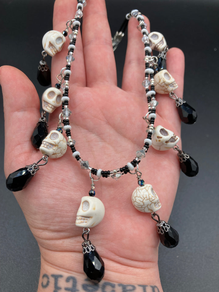 This statement necklace radiates a dark, shimmering aesthetic with two strands of glass beads connected through double-hole silver and black beads to create a collar shape. Three dangling skulls surrounded by black teardrop glass beads enable the piece to flow elegantly and express a bold, fierce look.