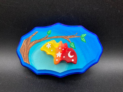 This unique Starry Night Mini Bat Wall Art Decor is hand sculpted and painted in vivid hues to offer an eye-catching decoration for your living space. The bat is painted in a gradient of yellow, to orange, to dark red, and is complemented by white stars and a crescent moon. The wood frame is painted in light to dark blues for a stunning night effect for a drastic contrast to the warm tone bat.