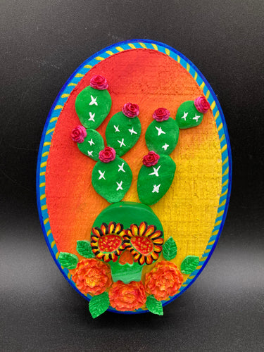 This handcrafted polymer clay wall art features a wood frame painted in a gradient from light yellow to dark red, creating a stunning sunset look. With a bright green skull cactus plant and bright pink blooming flowers. Boasting vivid marigold blooms and a deep royal blue frame to make a bold contrast with the warm background hue. this piece is sure to make a statement.  Measurements: H 7