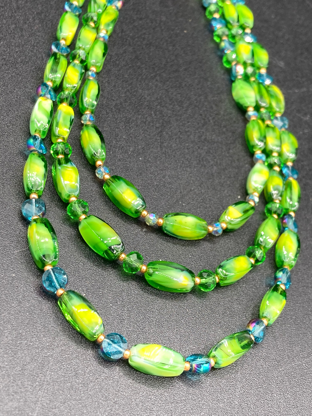 This adjustable glass beaded necklace features a combination of vivid green, blue, and gold hues and a mixture of shapes, creating an eye-catching spectrum of colors and a complex look. The rectangular green glass beads form the predominant part of the three-strand piece, which offers a bold and elegant fashion statement that is ideal for the Spring season.  Measurements:    Necklace is 17” in length, can be adjusted up to 19