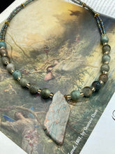 Load image into Gallery viewer, This exquisite necklace showcases earthy tones of mossy green ceramic beads, complemented by a gray toned howlite stone chip pendant. To add a sparkle, I interspersed gold-tone glass beads between each ceramic one. Two delicate strands feature small glass beads with hues of blue, gold, and purple. These are bridged by flower-shaped gold plated beads, which then unite with the round ceramic beads. This alluring earth-tone combination makes a stunning accessory. Necklace is 17” 
