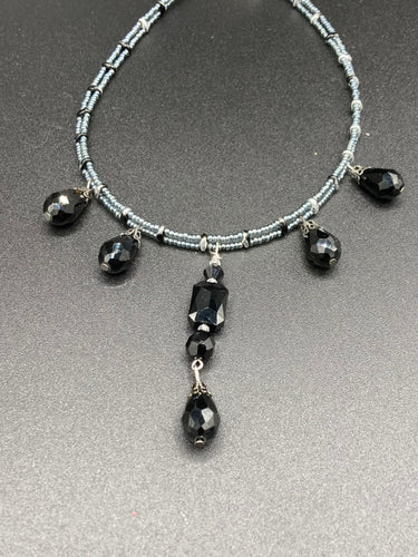This adjustable collar-style necklace features silver and black glass beads in a combination of both teardrop and rectangular shapes, designed to create visual complexity and depth. The two strands are held together with two-hole silver and black beads, creating a stunningly bold and elegant draped collar that enhances any outfit.