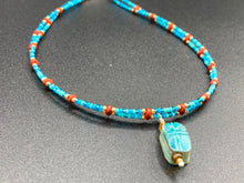Load image into Gallery viewer, This adjustable choker style necklace features an eye-catching teal wood scarab beetle pendant and glass beaded strands. This dramatic necklace is kept together by double-hole flat matte lava-red beads, providing a collar shape. The necklace artfully wraps around the neck, conveying an elegant and bold style.  Measurements:    Necklace is 14 1/2” in length, can be adjusted up to 16&quot; 
