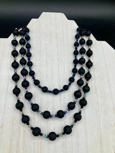 This statement piece features 3 long strands of flat matte black and dark blue glass beads, complemented by a floral metal beaded necklace. It gracefully drapes across the neck for a fierce, yet elegant look, making it perfect for cocktail chic attire, weddings, or minimalist styling.  Measurements:    Necklace is 16” in length - adjustable to 18